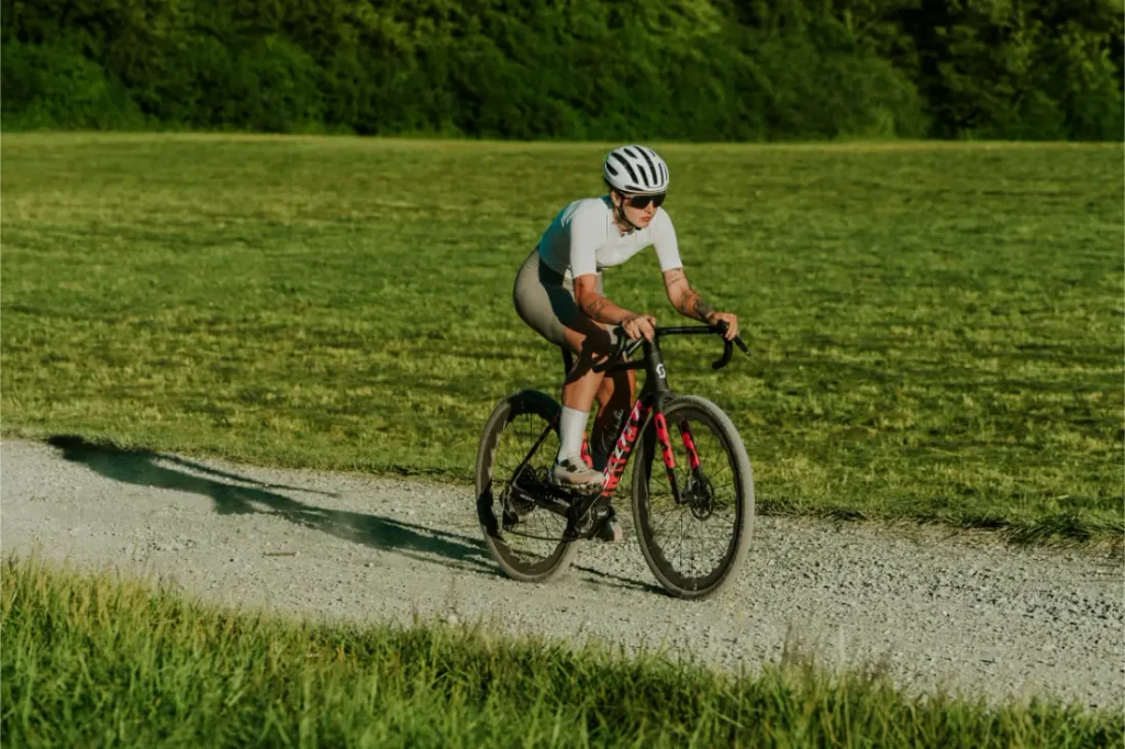 Un ciclista que lleva un casco blanco y ropa ligera de ciclismo va en bicicleta por un camino de grava que atraviesa un prado, con árboles al fondo.
