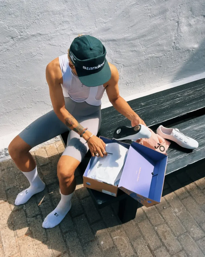 Una persona vestida con ropa de ciclista está sentada en un banco y abre una caja de zapatos en la que hay unas zapatillas blancas de ciclismo; una de las zapatillas está sobre el banco.