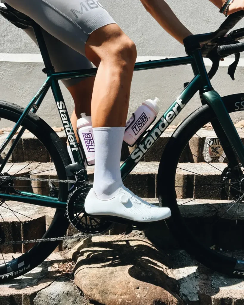 Una persona vestida con ropa de ciclista blanca está sentada en una bicicleta Stander verde con dos botellas de agua moradas sujetas al cuadro, situada en unos escalones de piedra.