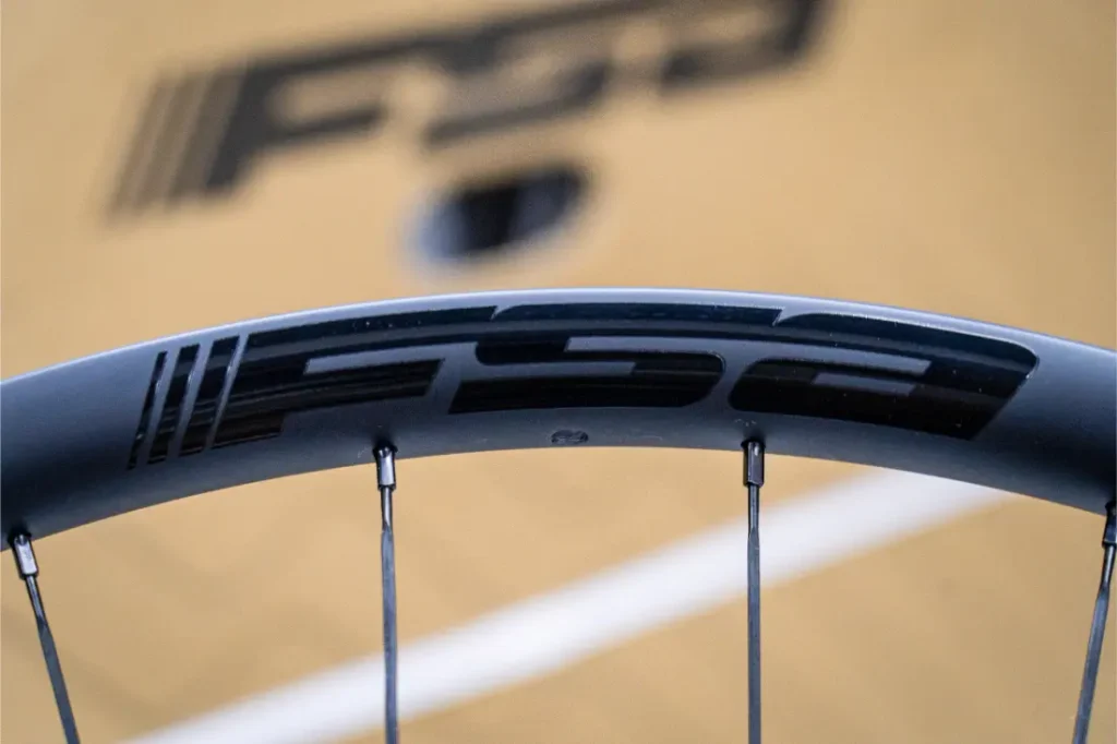 Primer plano de una llanta de bicicleta Ruedas FSA con la marca KFX i30, en el que se aprecian los radios y parte del logotipo sobre un fondo de color beige, lo que pone de relieve el control absoluto en cada detalle.