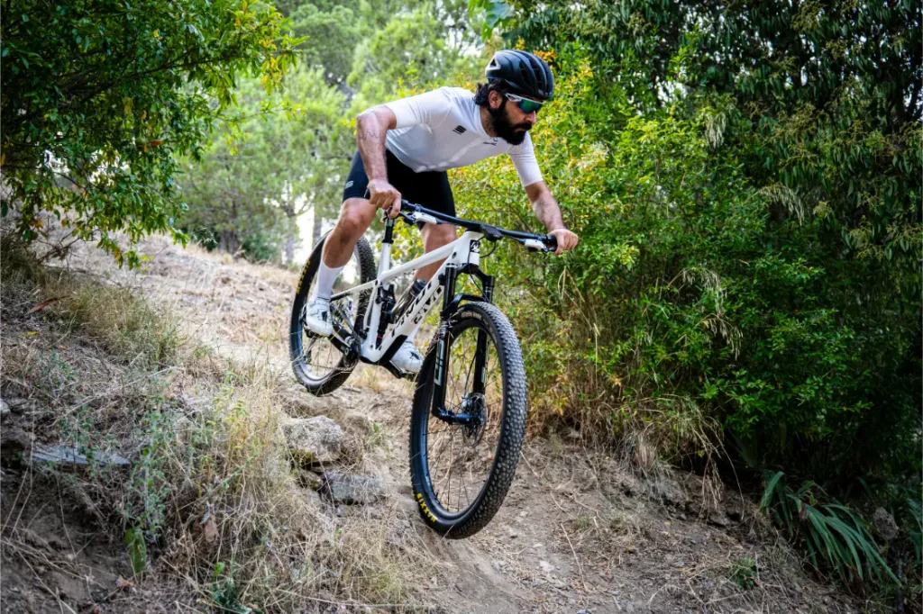 Una persona que lleva casco y gafas de sol desciende en una bicicleta de montaña KFX i30 por un sendero de tierra rodeado de árboles y arbustos, manteniendo con seguridad el control total sobre el terreno accidentado.