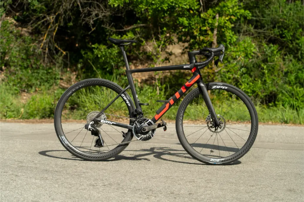Una bicicleta de carretera negra y roja, con manillar de carretera y ruedas Zipp, está aparcada en la acera; lleva neumáticos Schwalbe Pro One Aero montados en la rueda delantera y la rueda trasera, con vegetación y arbustos al fondo.
