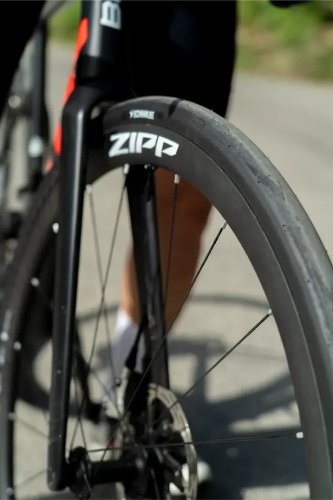 Primer plano de la rueda delantera de una bicicleta de carretera con la marca «Zipp», equipada con un neumático Schwalbe Pro One Aero, en el que se aprecia parte de la horquilla y el disco de freno sobre una superficie asfaltada.