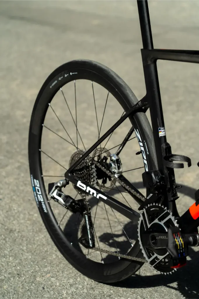 Primer plano de la rueda trasera, el cassette y la transmisión de una bicicleta de carretera negra equipada con un neumático Schwalbe Pro One sobre una superficie de asfalto.