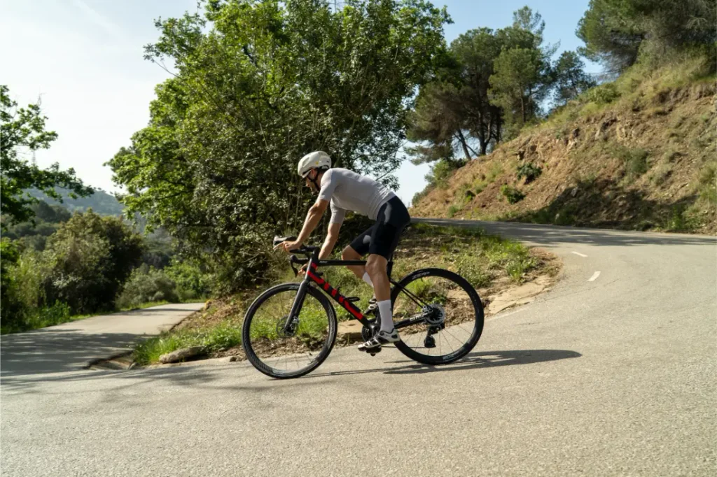 Un ciclista, que lleva casco y ropa deportiva, sube en bicicleta por una carretera sinuosa rodeada de árboles y vegetación, equipada con una rueda Aero Front y neumáticos Schwalbe Pro One para un mejor rendimiento.