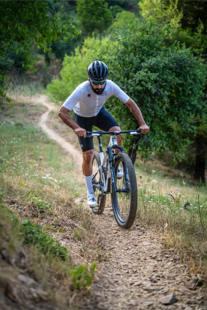 Un ciclista, que lleva casco y gafas de sol, pedalea en una bicicleta de montaña equipada con ruedas FSA cuesta arriba por un estrecho sendero de tierra rodeado de vegetación, demostrando un control total sobre el exigente terreno.