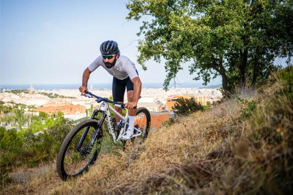 Una persona que lleva casco y gafas de sol pedalea cuesta arriba en una bicicleta de montaña equipada con ruedas FSA por un sendero cubierto de hierba, disfrutando de un control total mientras la ciudad y el mar se extienden al fondo.