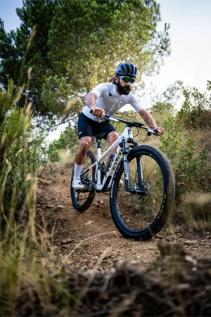 Una persona que lleva casco, gafas de sol y equipación de ciclismo circula en una bicicleta de montaña con ruedas FSA KFX i30 por un sendero de tierra rodeado de hierba y árboles, disfrutando de un control total sobre el terreno accidentado.