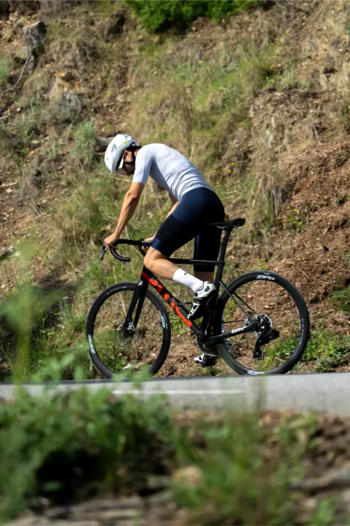 Un ciclista que lleva un casco blanco y un maillot gris pedalea cuesta arriba en una bicicleta de carretera negra, equipada con una rueda trasera Schwalbe Pro One, por una carretera asfaltada bordeada de césped y tierra.