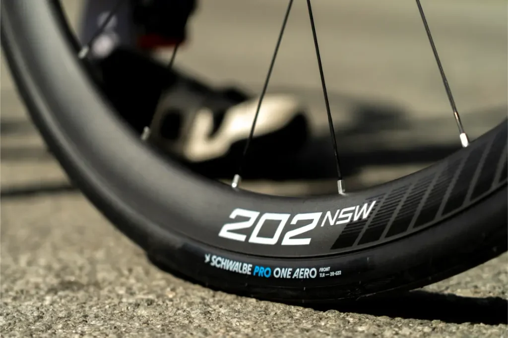 Primer plano de la rueda delantera de una bicicleta en el que se ven las inscripciones «202 NSW» y «Schwalbe Pro One Aero» en la llanta y el neumático, con parte de una zapatilla de ciclismo al fondo sobre una superficie pavimentada.