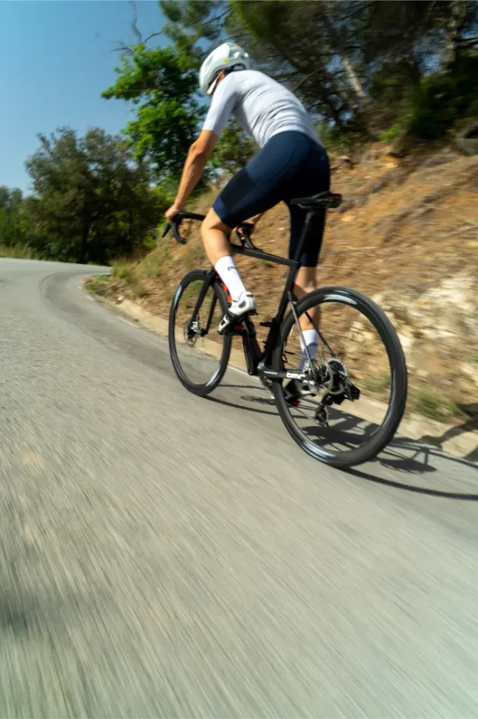Un ciclista, con casco blanco y ropa azul, pedalea cuesta arriba en una bicicleta de carretera equipada con neumáticos Schwalbe Pro One en un día soleado, con árboles y un camino en pendiente al fondo.