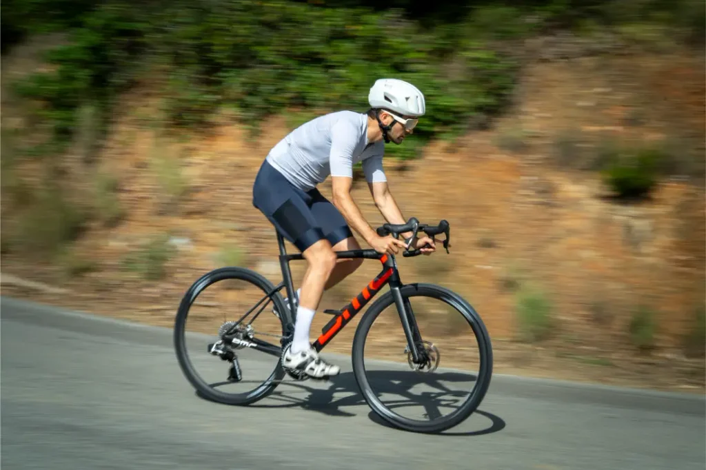 Un ciclista que lleva un casco blanco y un maillot gris pedalea en una bicicleta de carretera negra y roja equipada con neumáticos Schwalbe Pro One Aero tanto en la rueda delantera como en la trasera, deslizándose por un camino asfaltado con vegetación al fondo.