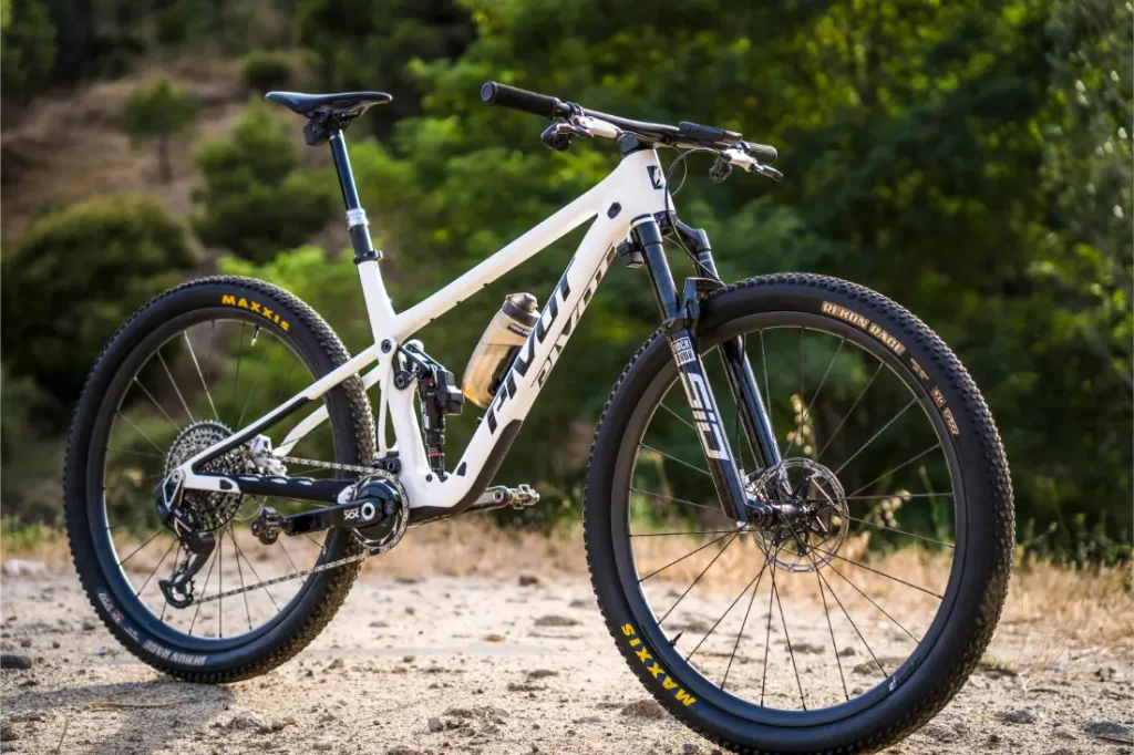 Una bicicleta de montaña blanca con suspensión total, equipada con neumáticos Maxxis y ruedas FSA KFX i30, está aparcada en un camino de tierra rodeado de árboles frondosos, lo que ofrece a los ciclistas un control total incluso en los terrenos más exigentes.
