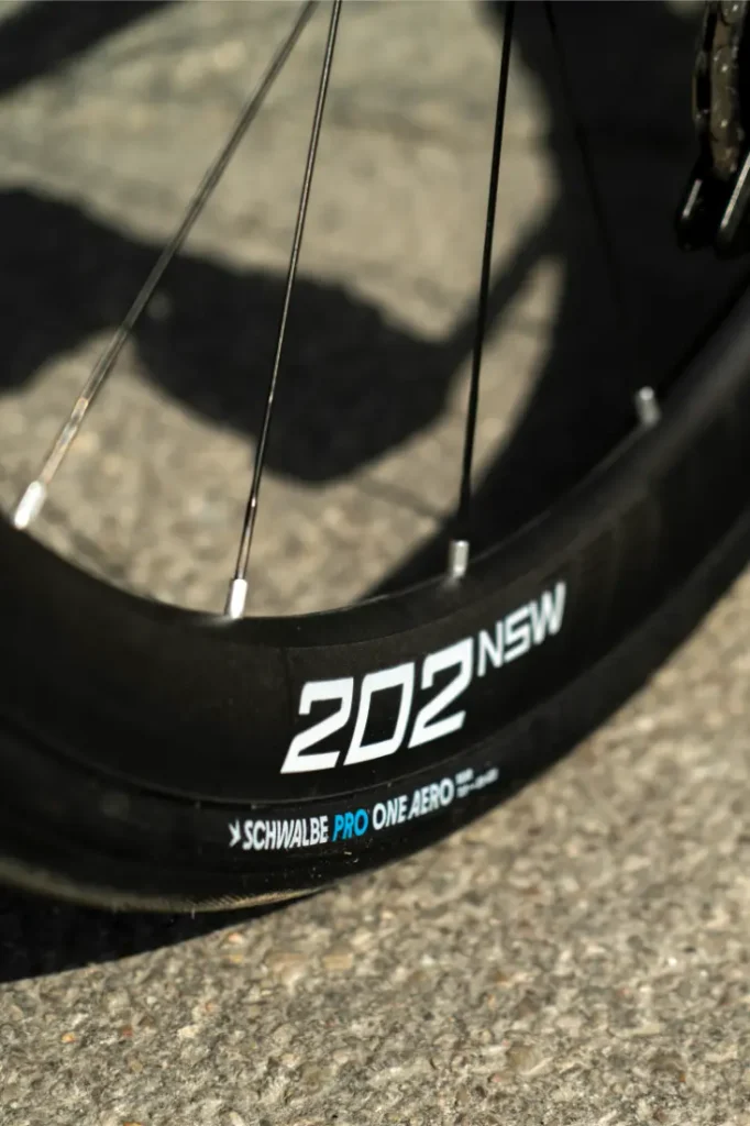 Primer plano de una rueda de bicicleta Aero Front en la que se aprecian las etiquetas «202 NSW» y «Schwalbe Pro One Aero» en la llanta, con el pavimento como fondo.