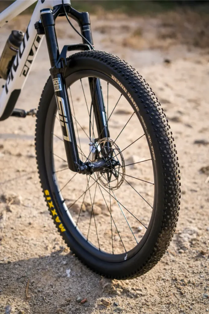 Primer plano de la rueda delantera y la horquilla de suspensión de una bicicleta de montaña, equipada con neumáticos Maxxis y ruedas FSA, en un sendero arenoso al aire libre para un control total.