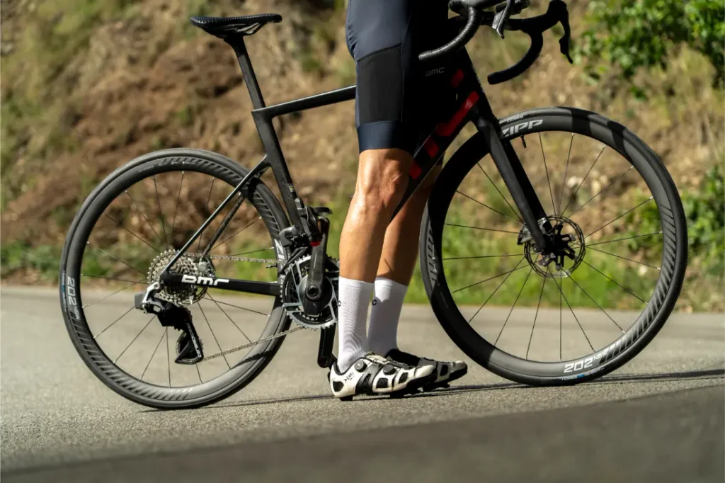 Un ciclista vestido de negro está de pie junto a una bicicleta de carretera negra equipada con neumáticos Schwalbe Pro One en una carretera asfaltada, con un pie en el suelo y el otro en un pedal.