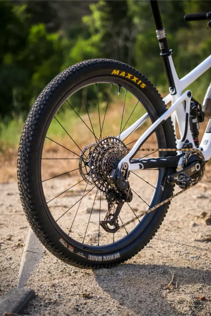 Primer plano de la rueda trasera y el sistema de cambios de una bicicleta de montaña equipada con ruedas FSA KFX i30 en un camino de tierra, con vegetación al fondo, que destaca el control total en cada recorrido.