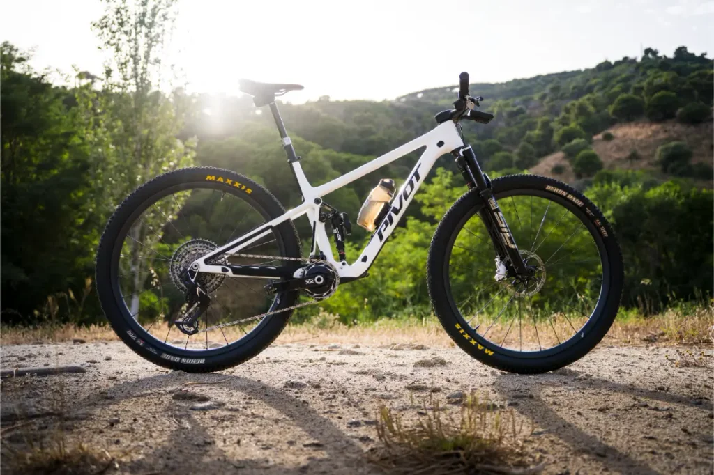 Una bicicleta de montaña blanca con suspensión total, equipada con ruedas FSA KFX i30 y neumáticos negros, está aparcada en un camino de tierra, ofreciendo un control total mientras descansa entre los árboles y las pintorescas colinas que se divisan al fondo.