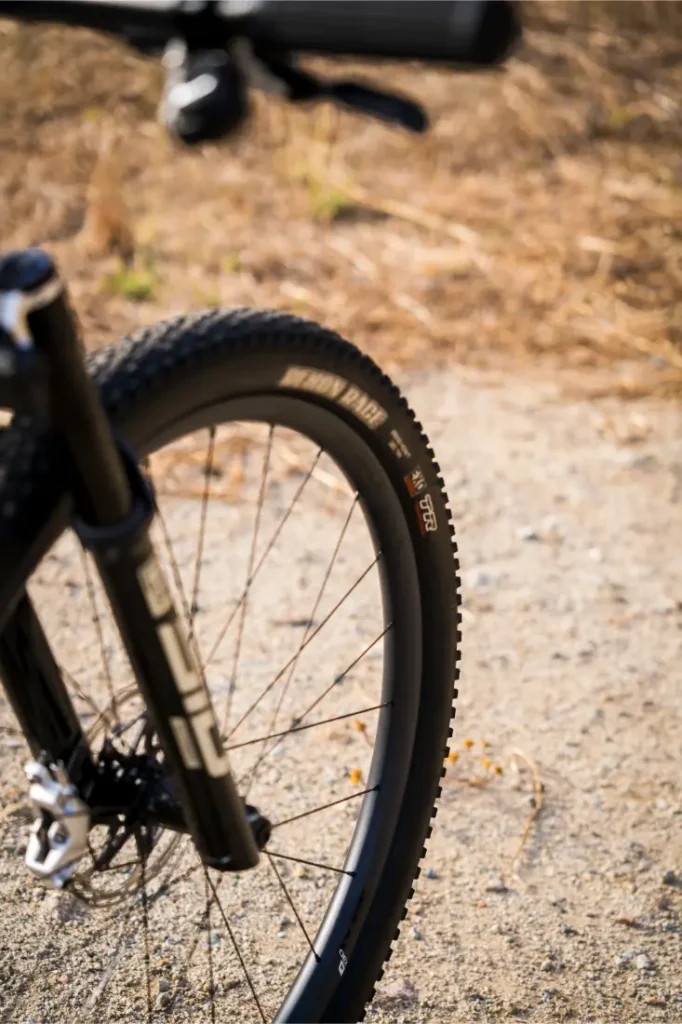 Primer plano de la rueda delantera y el neumático negros de una bicicleta de montaña en un camino de tierra, equipada con ruedas FSA para un control total, con hierba seca al fondo.