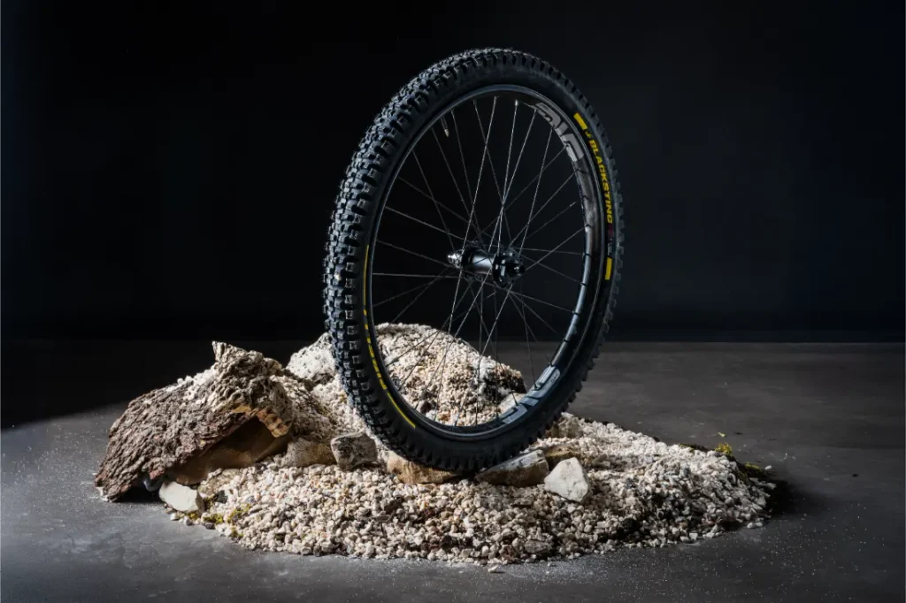 Una rueda de bicicleta de montaña Pirelli Scorpion se yace en posición vertical sobre un montón de rocas, grava y corteza, con un fondo oscuro, lo que pone de manifiesto la gran resistencia de los neumáticos de MTB.