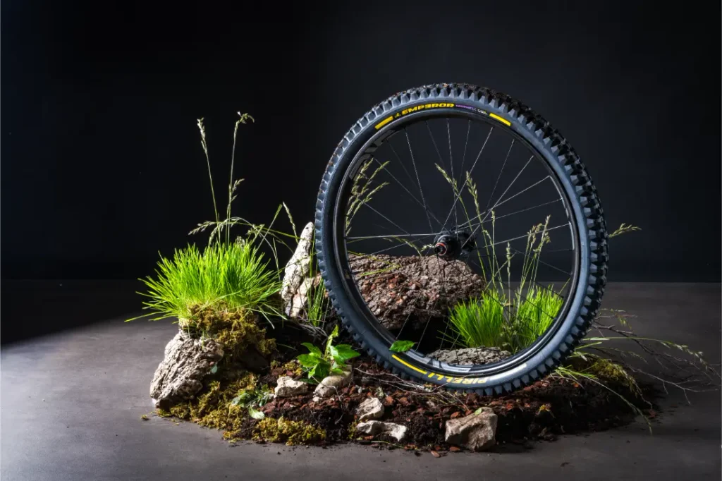 Una rueda de bicicleta de montaña equipada con neumáticos Pirelli Scorpion MTB aparece en posición vertical entre rocas, hierba y tierra, sobre un fondo oscuro.