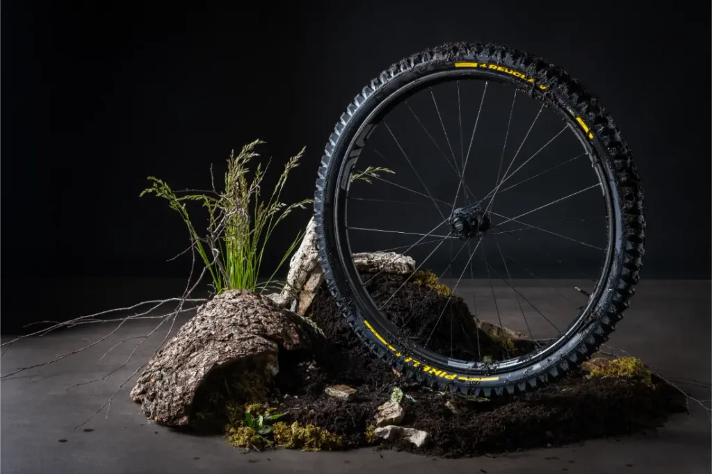 Una rueda de bicicleta de montaña con neumáticos Pirelli Scorpion MTB se yergue sobre tierra y musgo, rodeada de hierba y corteza, con un fondo oscuro como telón de fondo.