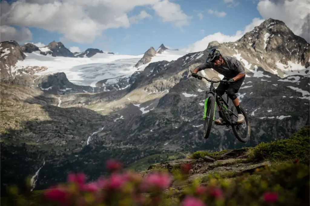 Una persona monta en bicicleta de montaña, equipada con neumáticos Pirelli Scorpion MTB, y se lanza desde un saliente rocoso, desafiando la gravedad con montañas nevadas y un glaciar al fondo, y flores silvestres de color rosa en primer plano.