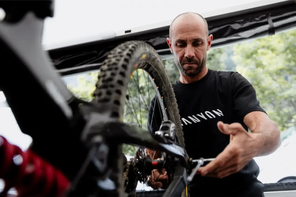 Un hombre que lleva una camiseta negra de Canyon repara una bicicleta de montaña, prestando especial atención a la cadena y los cambios. Equipada con unos robustos neumáticos Pirelli Scorpion para MTB, ideales para senderos de gravedad, su bicicleta destaca sobre el fondo de los árboles y parte de una copa.