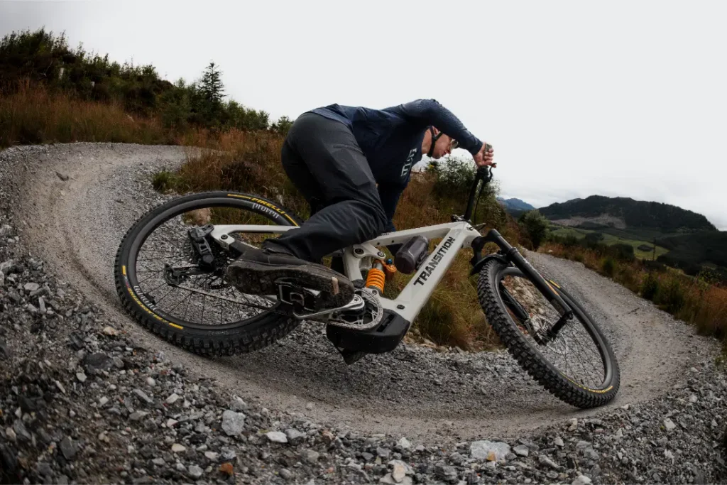 Una persona recorre una curva de grava en un sendero de tierra con una bicicleta de montaña Transition equipada con neumáticos Pirelli Scorpion MTB, rodeada de hierba y colinas lejanas bajo un cielo nublado.