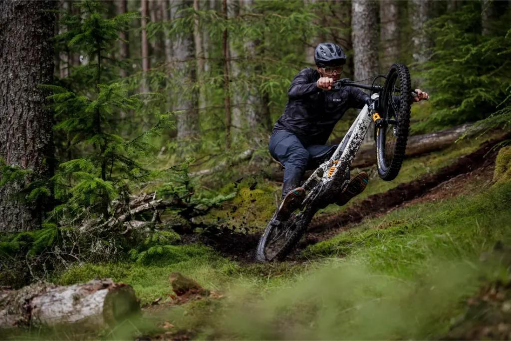 Una persona con casco monta en bicicleta de montaña equipada con neumáticos Pirelli Scorpion MTB por un sendero forestal embarrado, levantando tierra e inclinándose en las curvas entre árboles y vegetación.