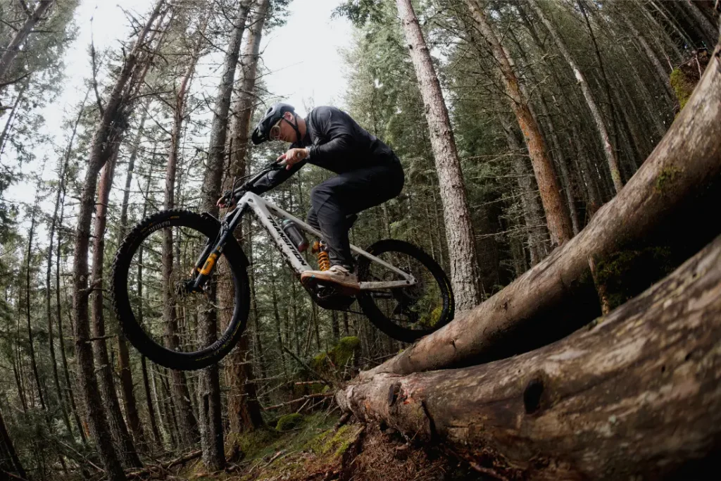 Una persona que lleva casco monta en una bicicleta de montaña equipada con neumáticos Pirelli Scorpion MTB y salta desde un tronco caído en un bosque frondoso; la imagen está tomada desde un ángulo bajo.
