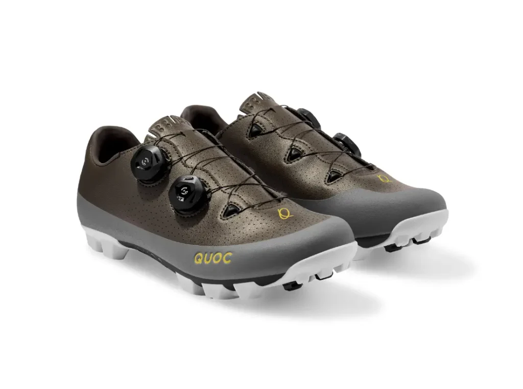 Un par de zapatillas de ciclismo marrones y grises con cierres de dial negros, suelas blancas y el logotipo «QUOC» en el lateral.