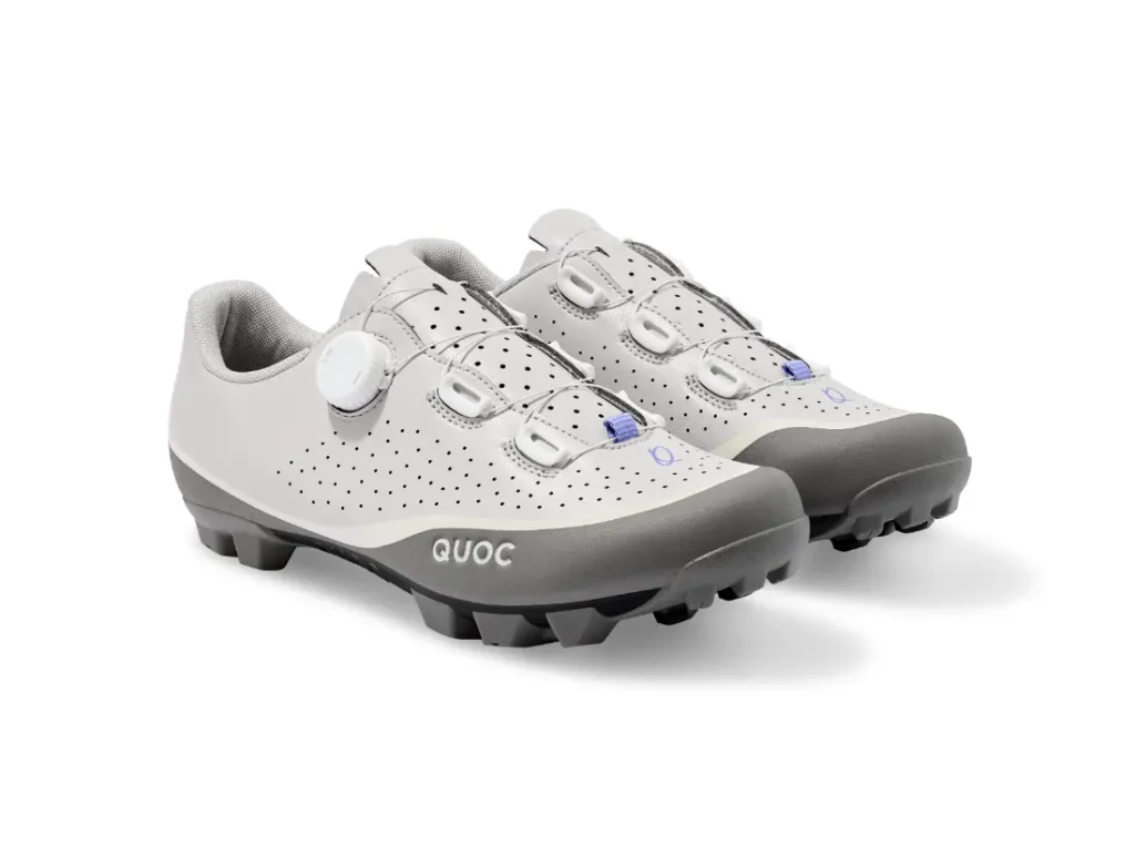 Un par de zapatillas de ciclismo blancas y grises con tacos negros, parte superior perforada y sistema de cierre con rueda giratoria.
