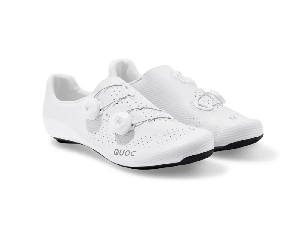 Un par de zapatillas de ciclismo blancas con suela negra, con la parte superior perforada y sistema de cierre con rueda giratoria, colocadas una al lado de la otra sobre un fondo blanco.