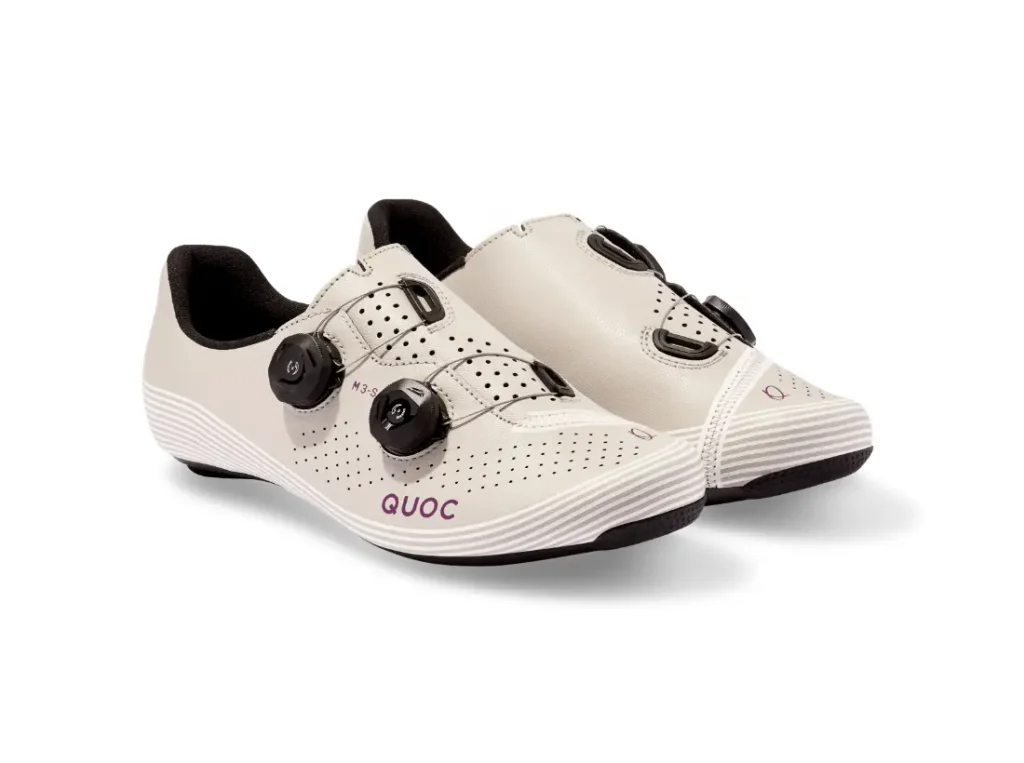 Un par de zapatillas de ciclismo de color beige con cierres negros, parte superior perforada y el nombre de la marca «QUOC» impreso en morado en el lateral.