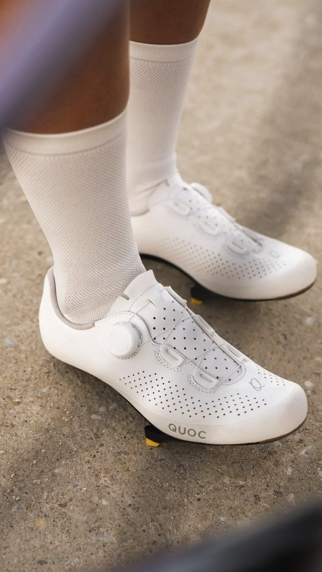Primer plano de una persona que lleva zapatillas de ciclismo blancas con perforaciones y calcetines blancos, de pie sobre una superficie de hormigón.