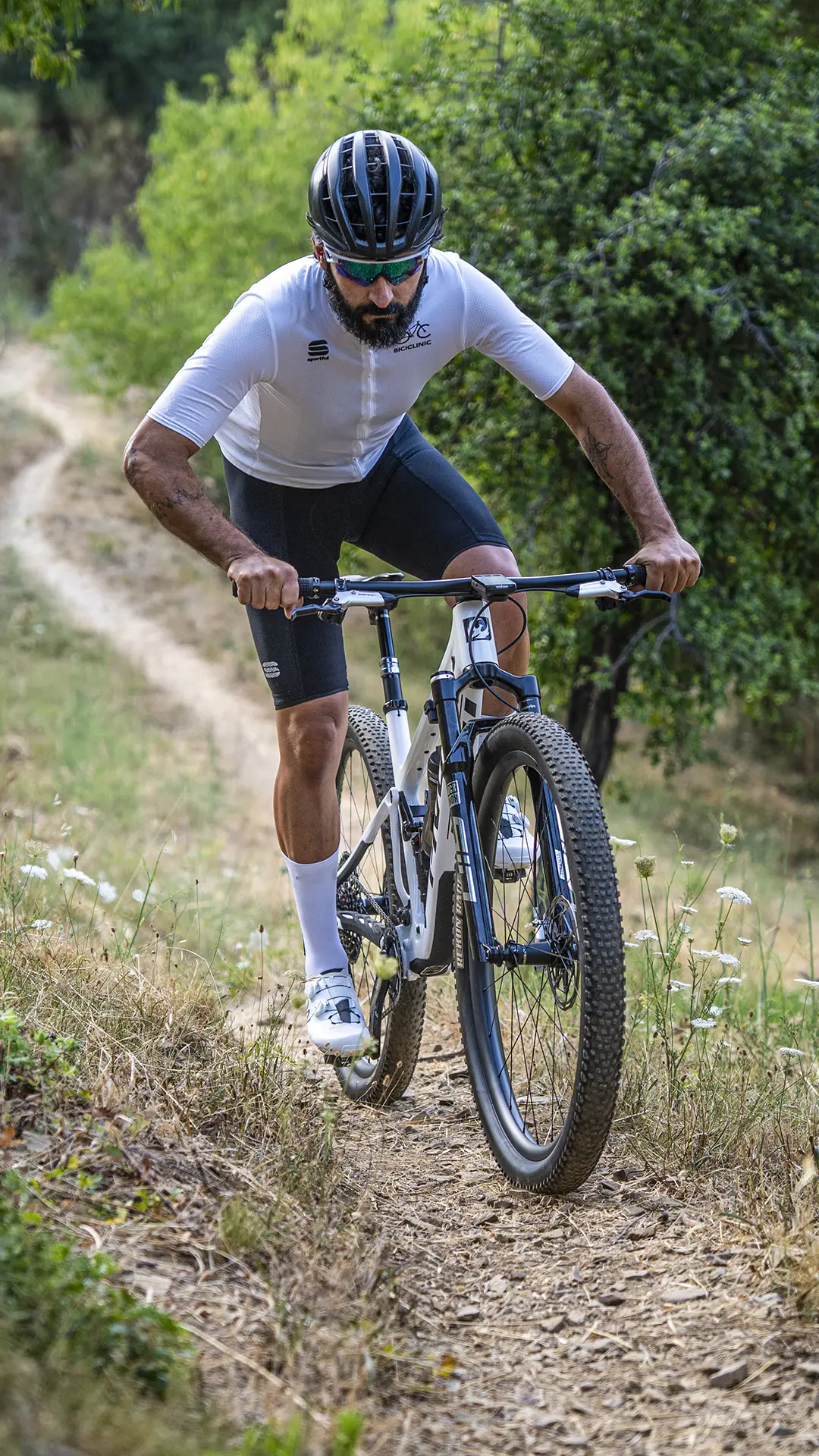 Un ciclista, que lleva casco y gafas de sol, sube en una bicicleta de montaña KFX i30 por un estrecho sendero de tierra que atraviesa una zona boscosa, manteniendo con destreza el control absoluto en este terreno tan exigente.