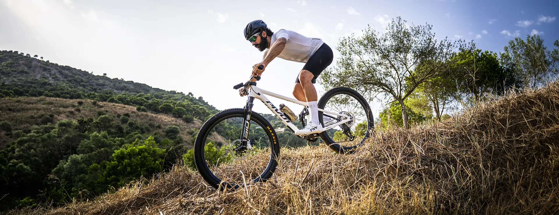 Una persona con casco baja en bicicleta de montaña, equipada con ruedas FSA KFX i30, por una colina seca y cubierta de hierba, demostrando un control total, con árboles y un cielo azul al fondo.