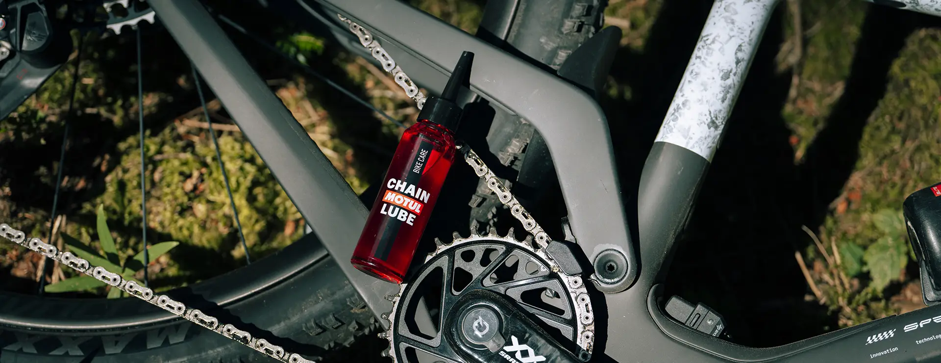 Una botella roja con la etiqueta «Chain Motul Lube» está colocada en el cuadro de una bicicleta de montaña, cerca de la cadena y el sistema de cambios, al aire libre.