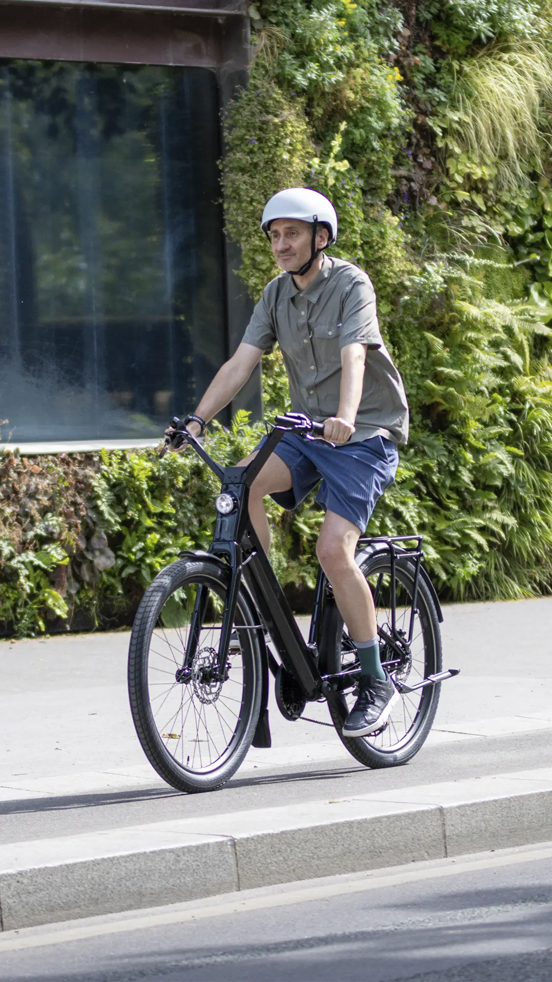 Un hombre con casco y ropa informal circula en una bicicleta eléctrica «Moustache Lundi 27» por una calle de la ciudad, con un muro verde de plantas al fondo, lo que refleja el espíritu vibrante de la ciudad.