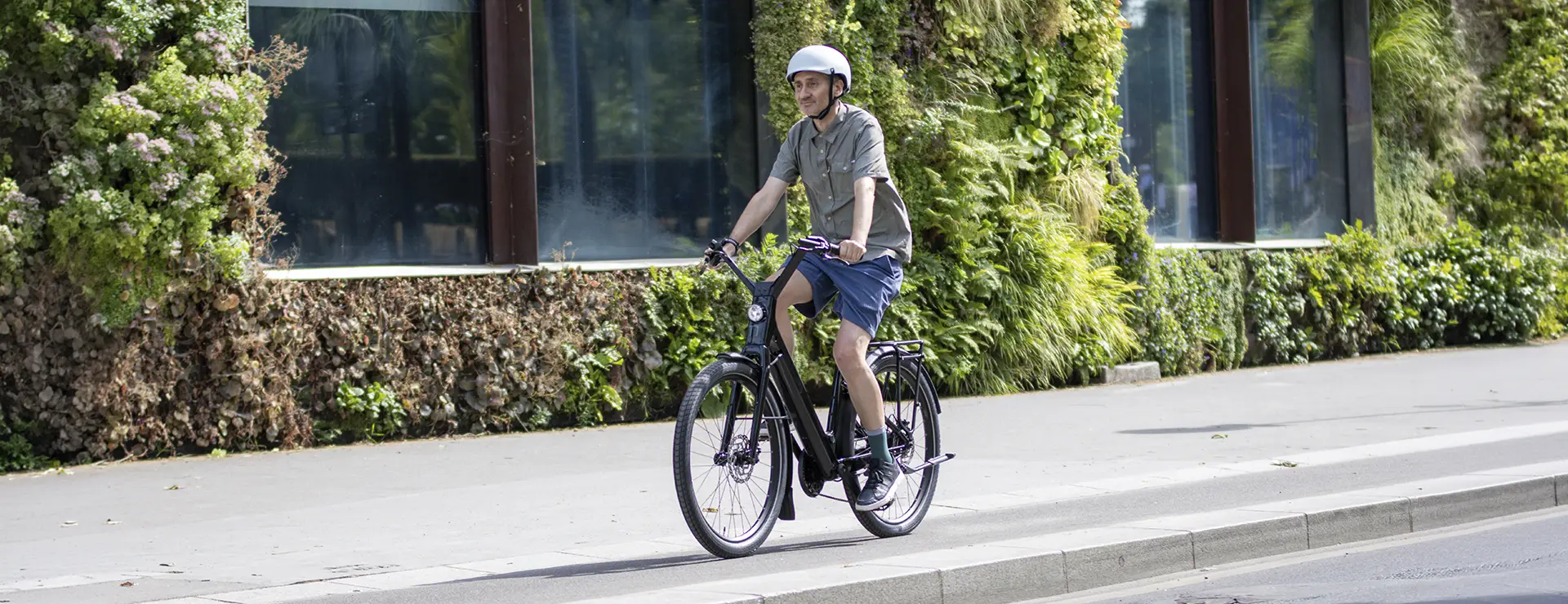 Una persona que lleva casco va en una bicicleta «Moustache Lundi 27» por una acera de la ciudad, junto a un edificio con plantas verdes en la fachada.