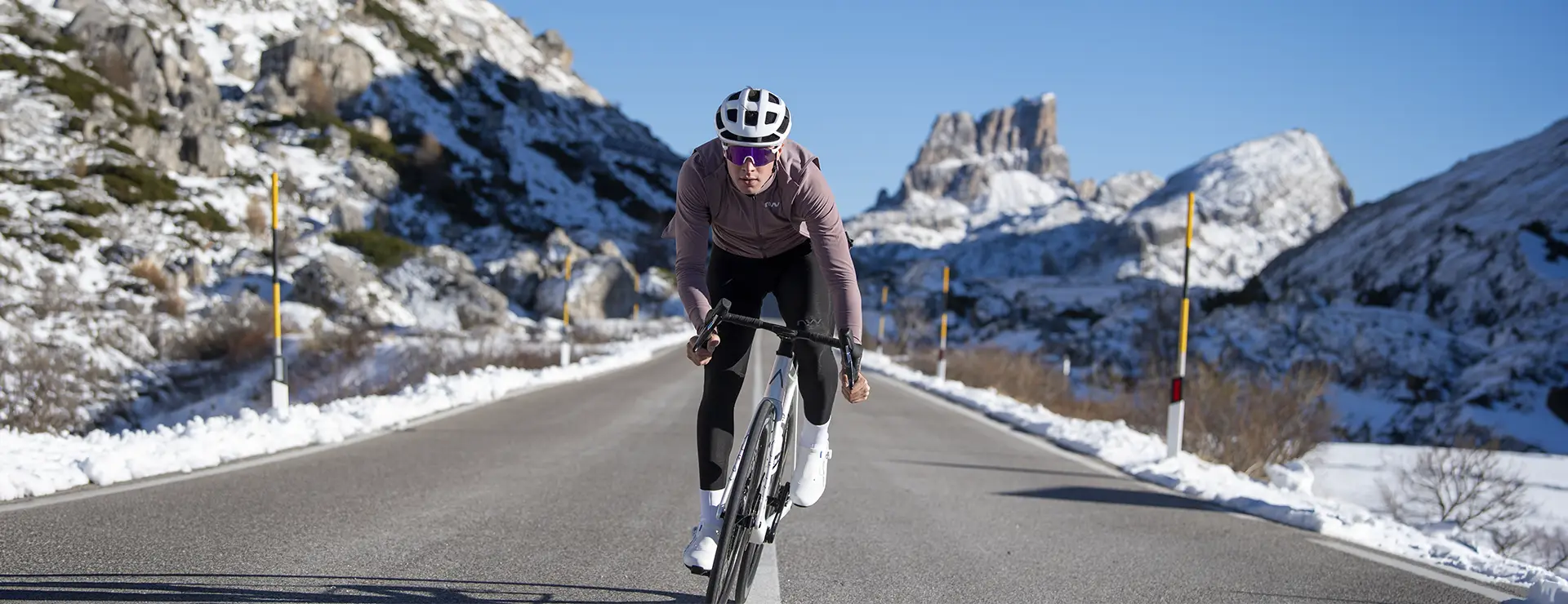 Un ciclista, con casco y gafas de sol, pedalea por una carretera de montaña asfaltada, bordeada de nieve, bajo un cielo azul y despejado.
