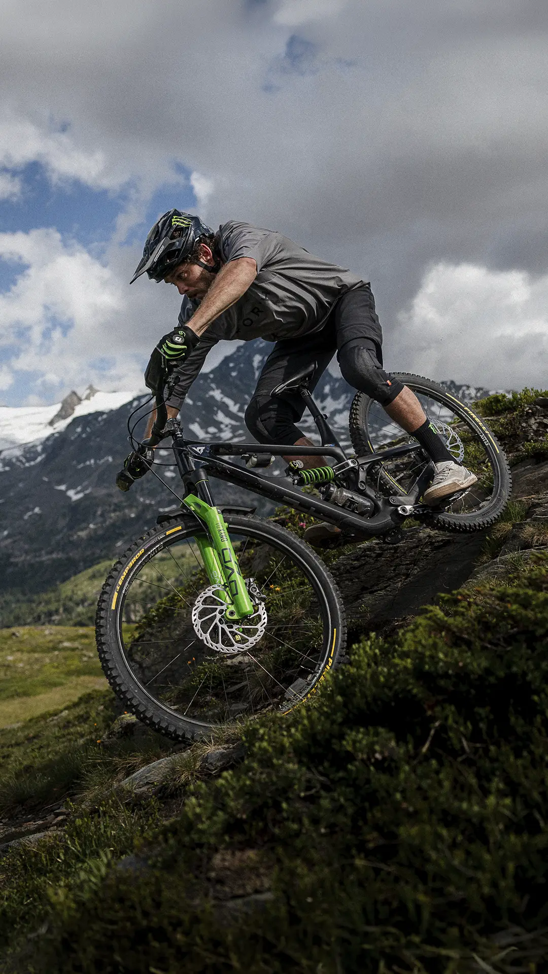 Un ciclista, que lleva casco y guantes, desciende en una bicicleta de montaña equipada con neumáticos Pirelli Scorpion MTB por una pendiente empinada y rocosa, con las montañas y las nubes como telón de fondo.