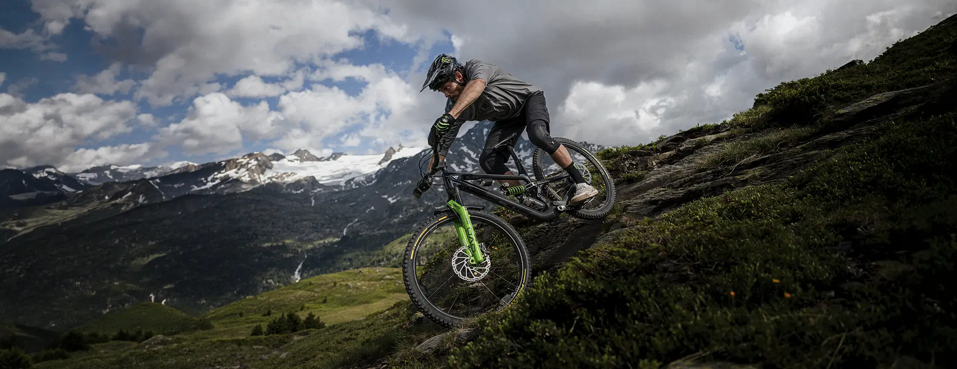 Un ciclista de montaña desciende por un sendero rocoso, equipado con neumáticos Pirelli Scorpion para MTB, rodeado de verdes colinas y lejanas montañas nevadas bajo un cielo nublado.