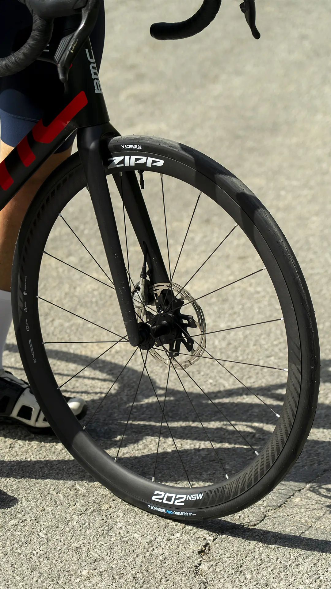 Primer plano de la rueda delantera de una bicicleta de carretera con llanta de carbono Zipp 202 NSW y freno de disco, equipada con un neumático Schwalbe Pro One Aero, colocada sobre una superficie asfaltada.