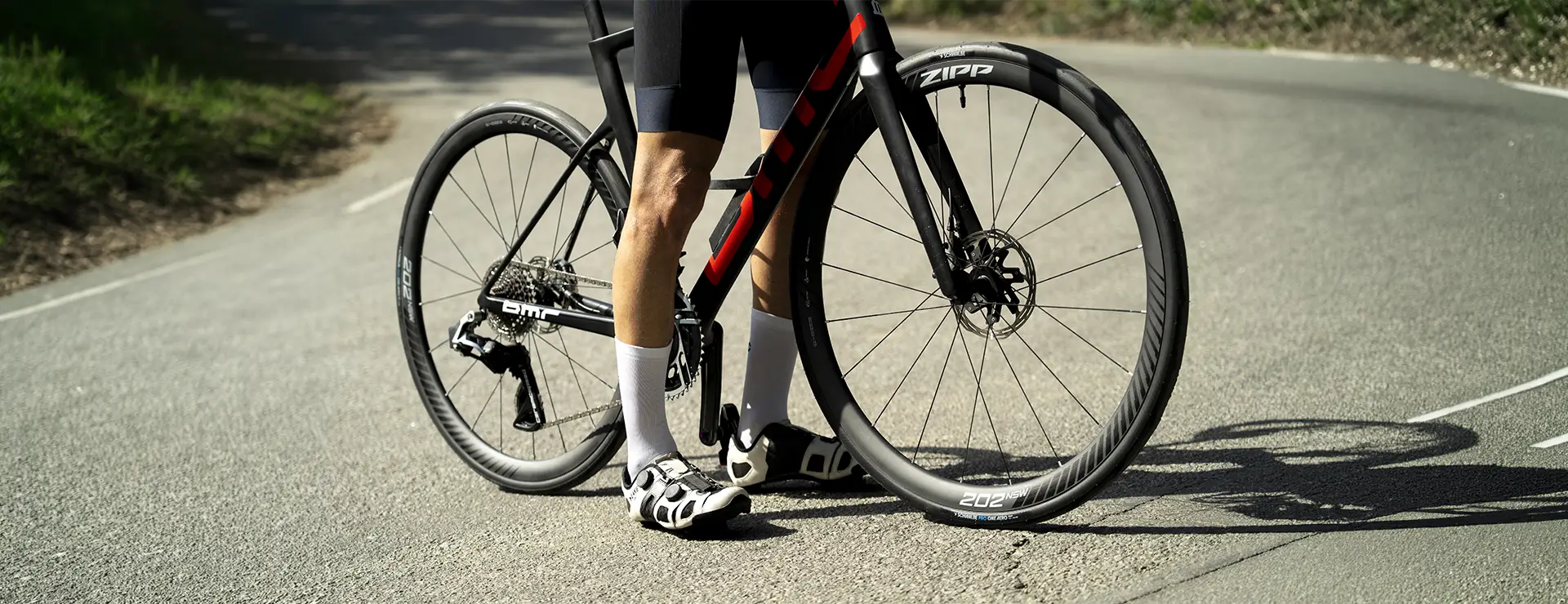 Un ciclista posa junto a una bicicleta de carretera con ruedas Zipp en una carretera asfaltada; la rueda delantera lleva un neumático Schwalbe Pro One Aero, y él lleva pantalones cortos negros, calcetines blancos y zapatillas de ciclismo.