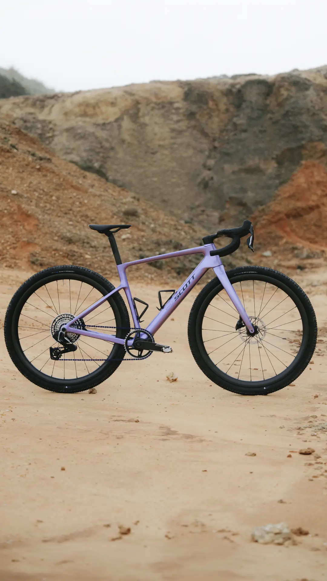 Una bicicleta gravel SCOTT Addict Gravel 2027 de color morado, con neumáticos negros, está aparcada sobre terreno arenoso, con un paisaje rocoso y montañoso al fondo.