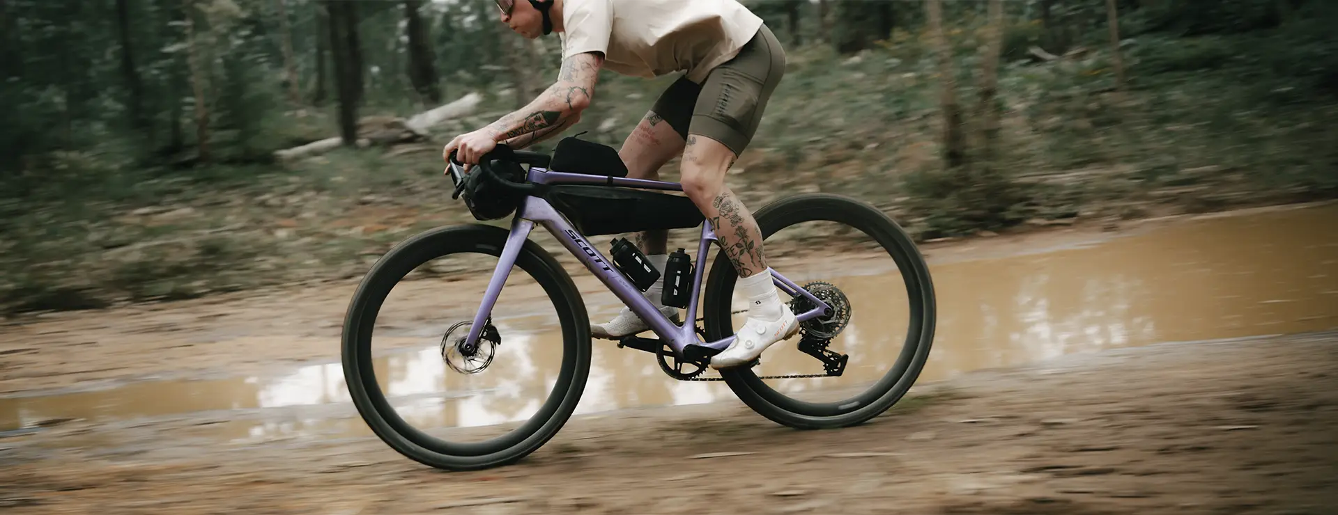 Una persona que va en una bicicleta SCOTT Addict Gravel 2027 de color morado por un sendero embarrado que atraviesa un bosque, vestida con pantalones cortos, una camiseta ligera y gafas de sol.