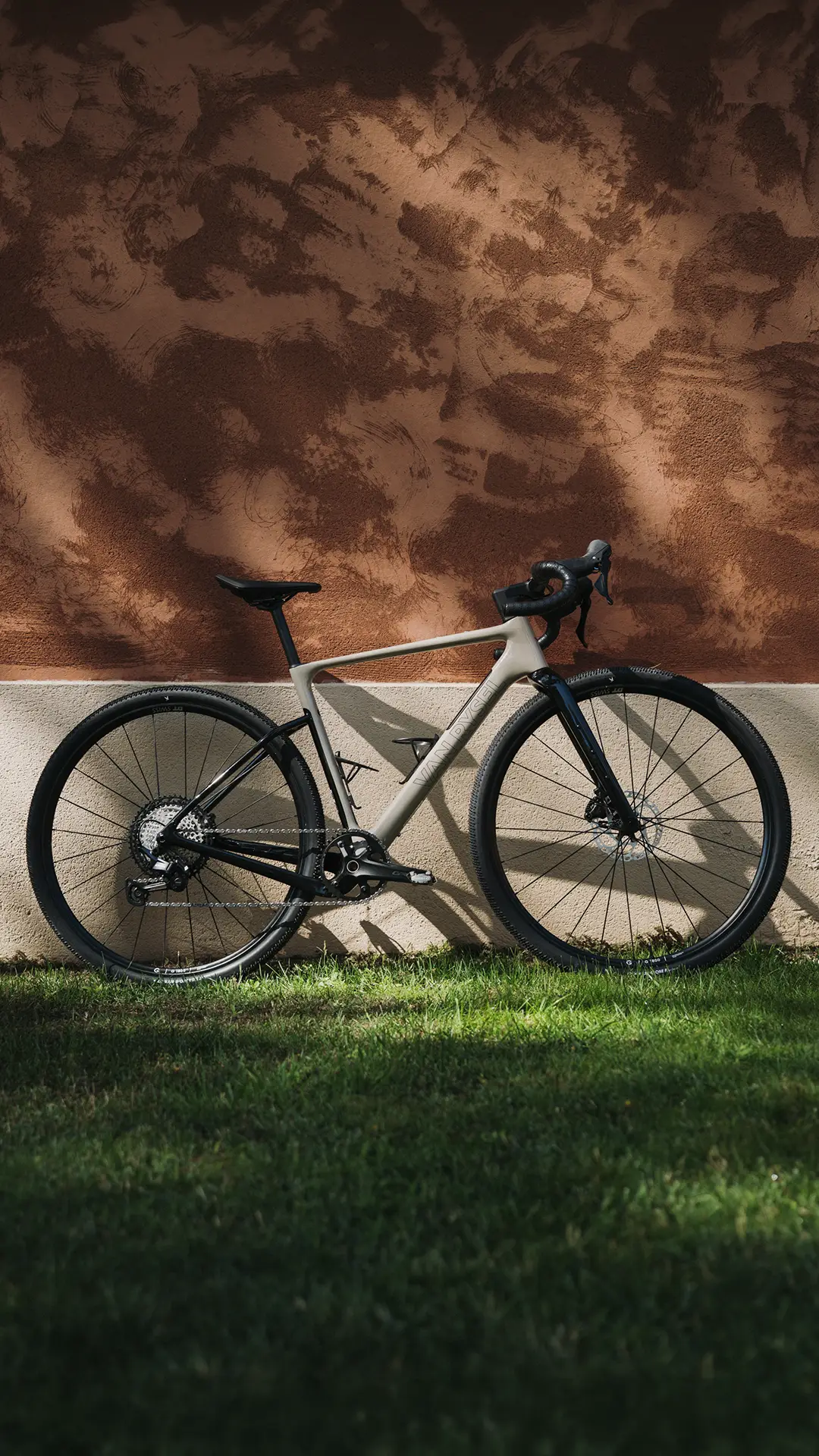 Una moderna bicicleta de carretera Van Rysel EDR-G con manillar de carretera está aparcada sobre la hierba verde, frente a una pared con textura en tonos marrones y blancos.
