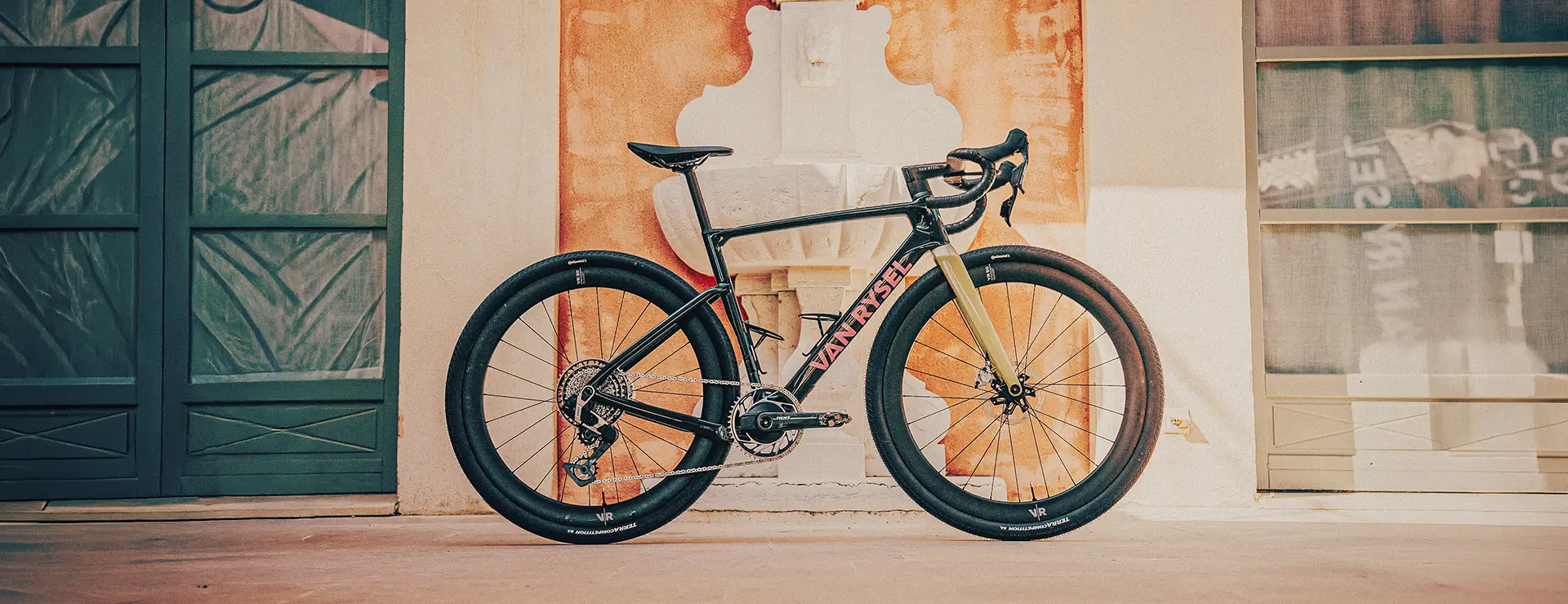 Una bicicleta de carretera Ventum, de color negro y rojo y con neumáticos anchos, está aparcada en posición vertical junto a un mural, cerca de una fuente y de unos grandes ventanales; su llamativo diseño recuerda a modelos de alto rendimiento como la Van Rysel RCR-G.